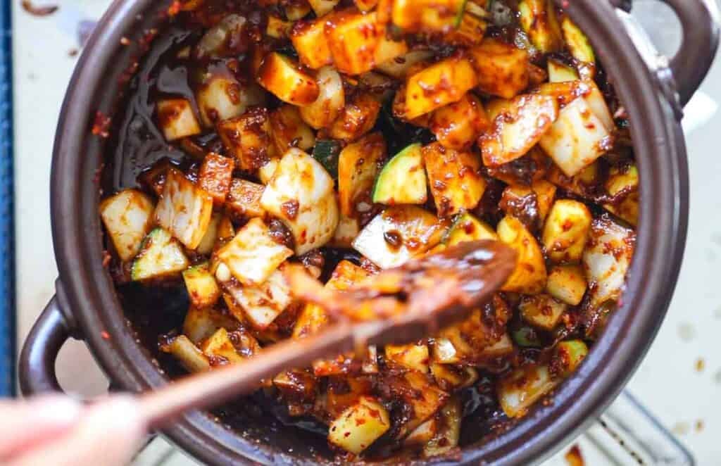 Close-up of a pot filled with diced vegetables, likely cucumbers and radishes, mixed with a spicy red sauce. A wooden spoon is seen stirring the ingredients, showcasing the vibrant and flavorful mixture.